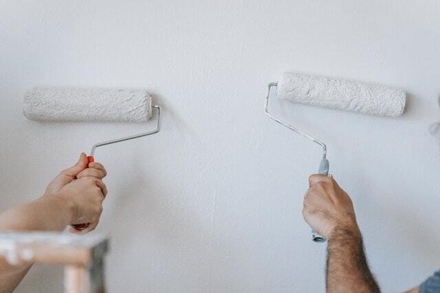 Two people's hands using paint rollers to paint a white wall